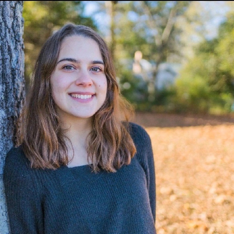 Person standing next to a tree, wearing a blue shirt with trees in the background.